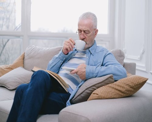 A mature man drinking coffee and looking relaxed while reading a book by the window